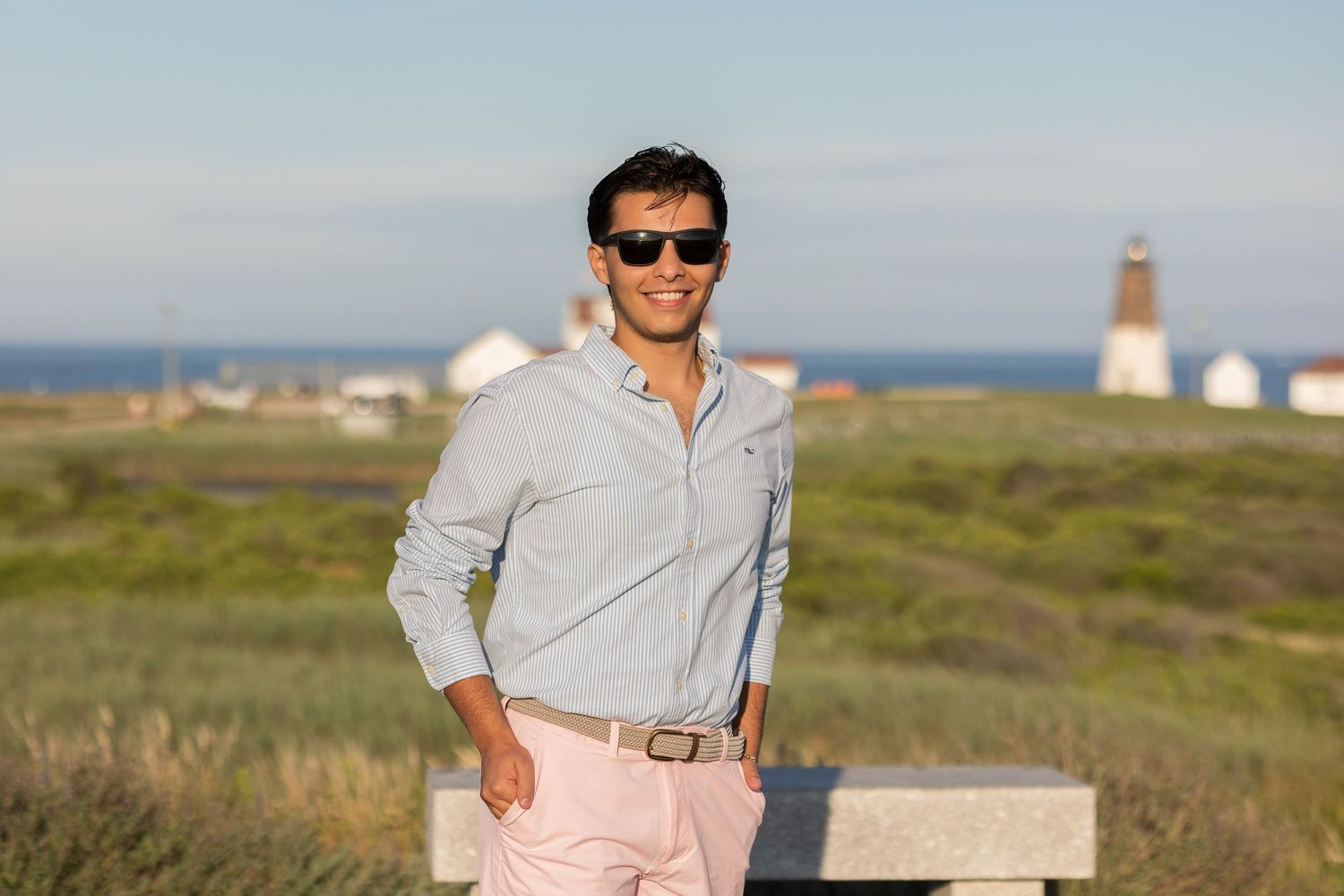A person in sunglasses and light colored clothing stands near a lighthouse on a grassy coastal landscape.