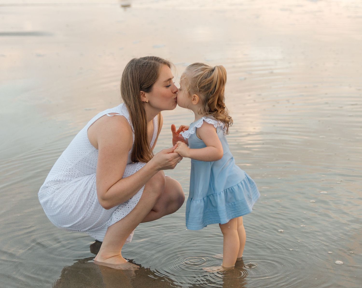 A tender waterside moment between parent and child in white and blue dresses at sunset.
