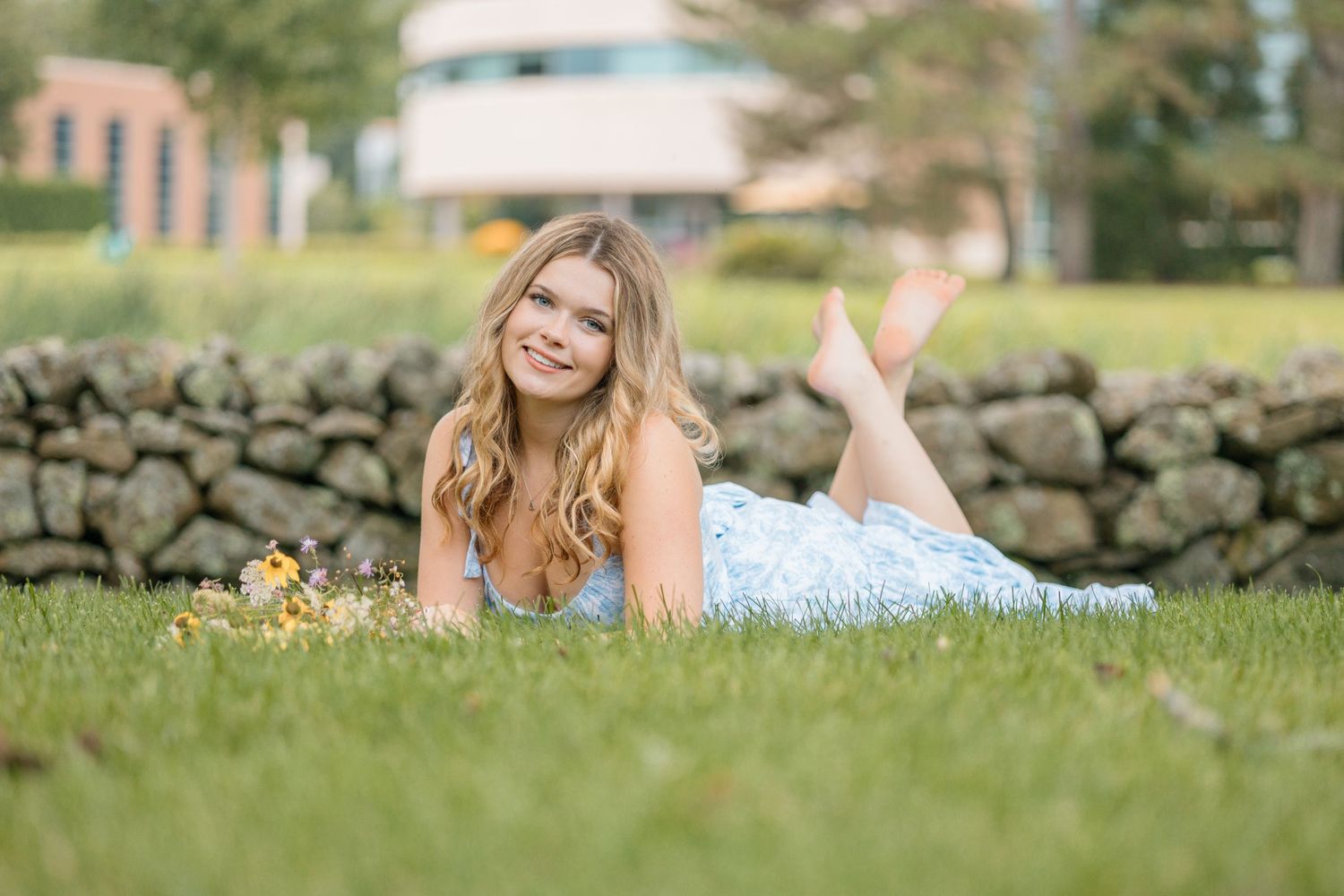 A person in a white dress lies on lush green grass near a stone wall on a sunny day.