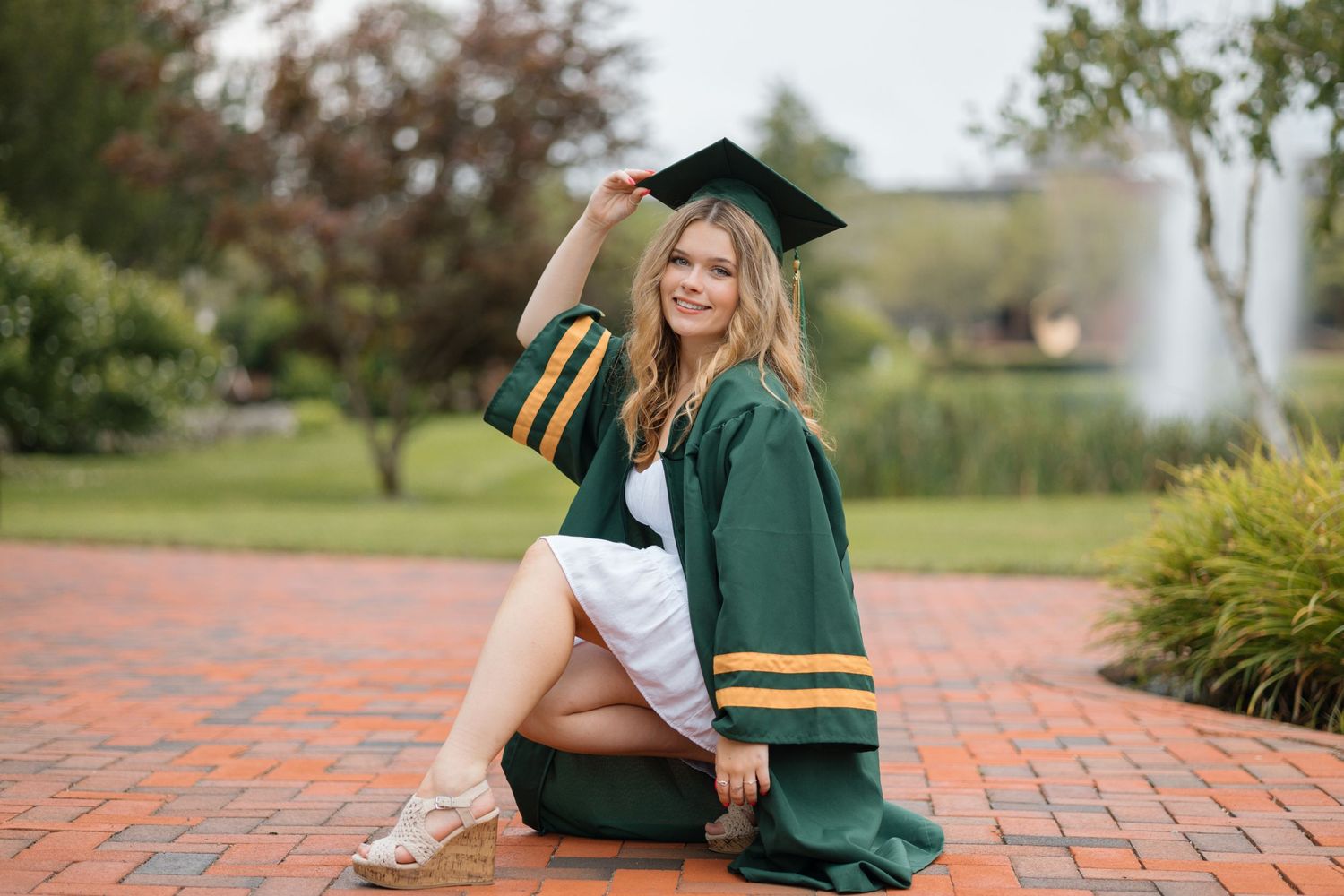 A graduate in green and gold academic regalia sits on brick pavement during commencement.