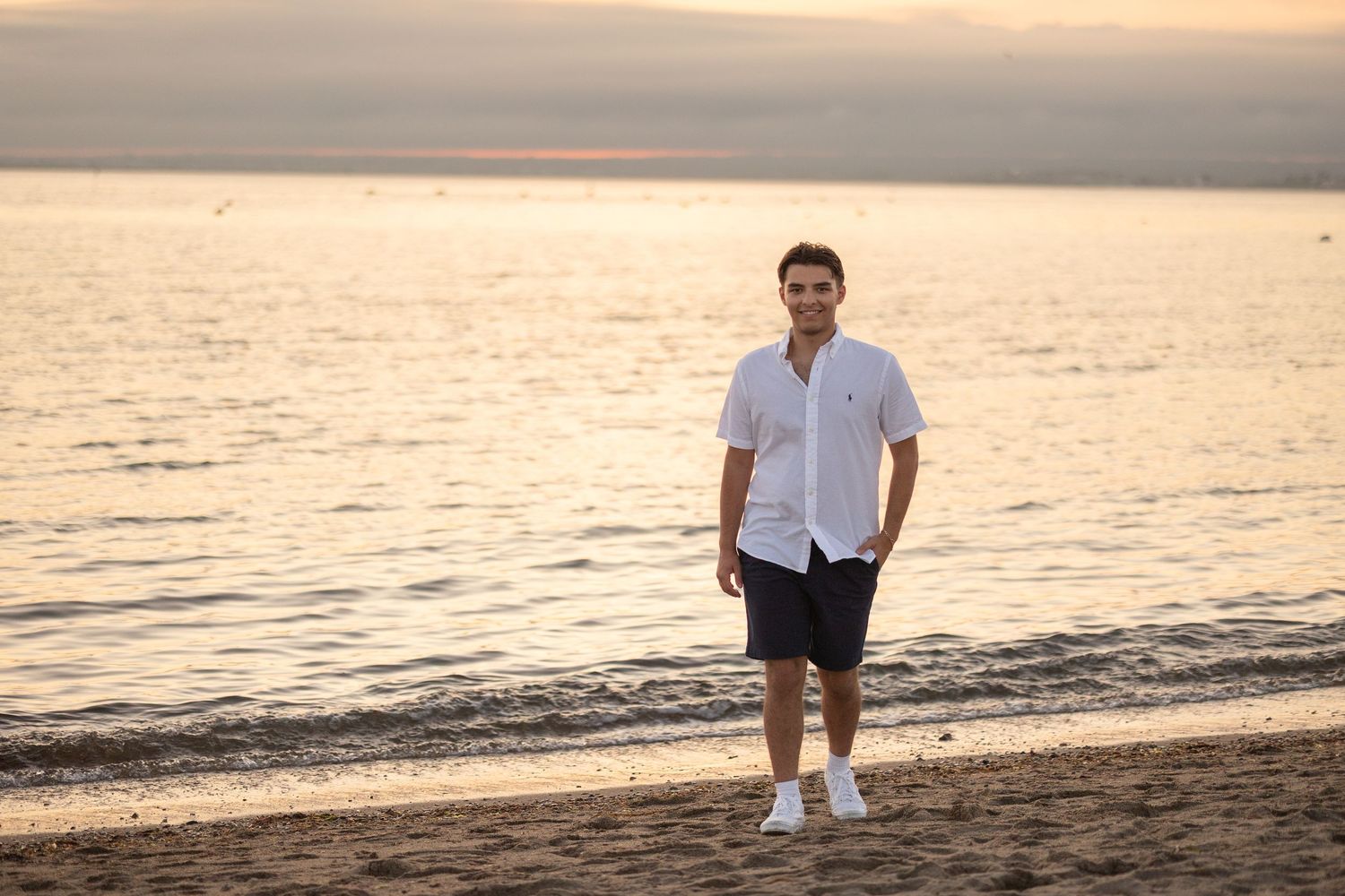 Person in white shirt and navy shorts stands on beach at sunset with calm ocean waters in background.