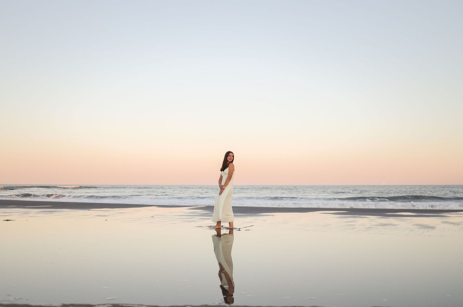 A silhouetted figure stands at the edge of a tranquil beach during sunset, creating a peaceful coastal scene.