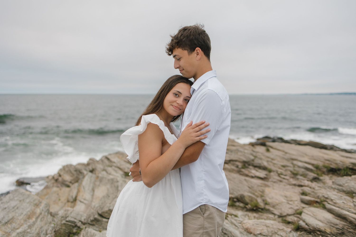 Couple in white embracing on rocky coastal shoreline with ocean waves and cloudy sky in background.