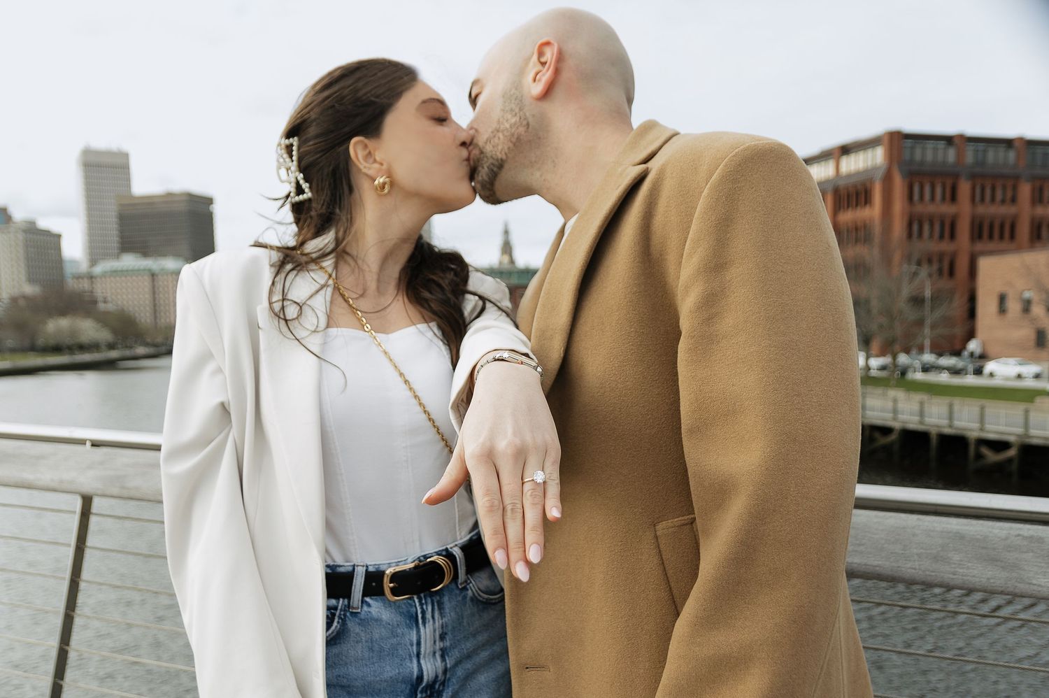 A romantic embrace on a waterfront deck with urban architecture in the background.