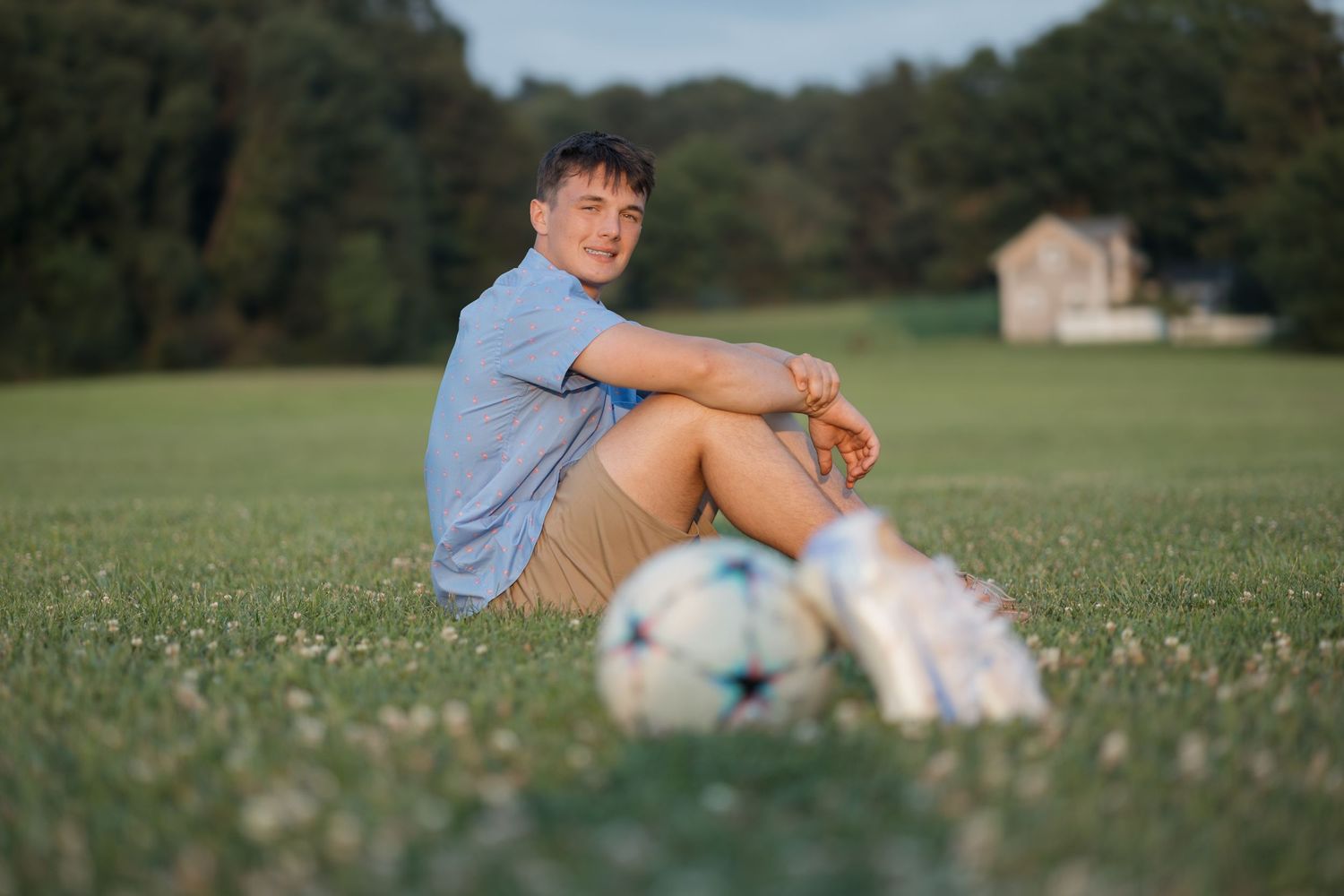 Person sitting in grassy field next to soccer ball with rural building in background.