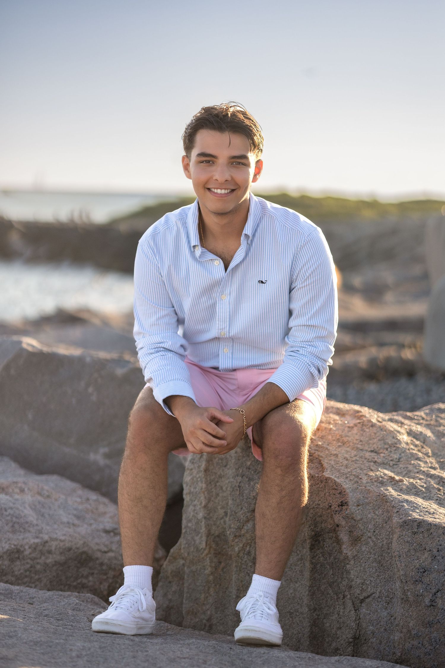 A person in a white button-down shirt and pink shorts sits on coastal rocks during golden hour.