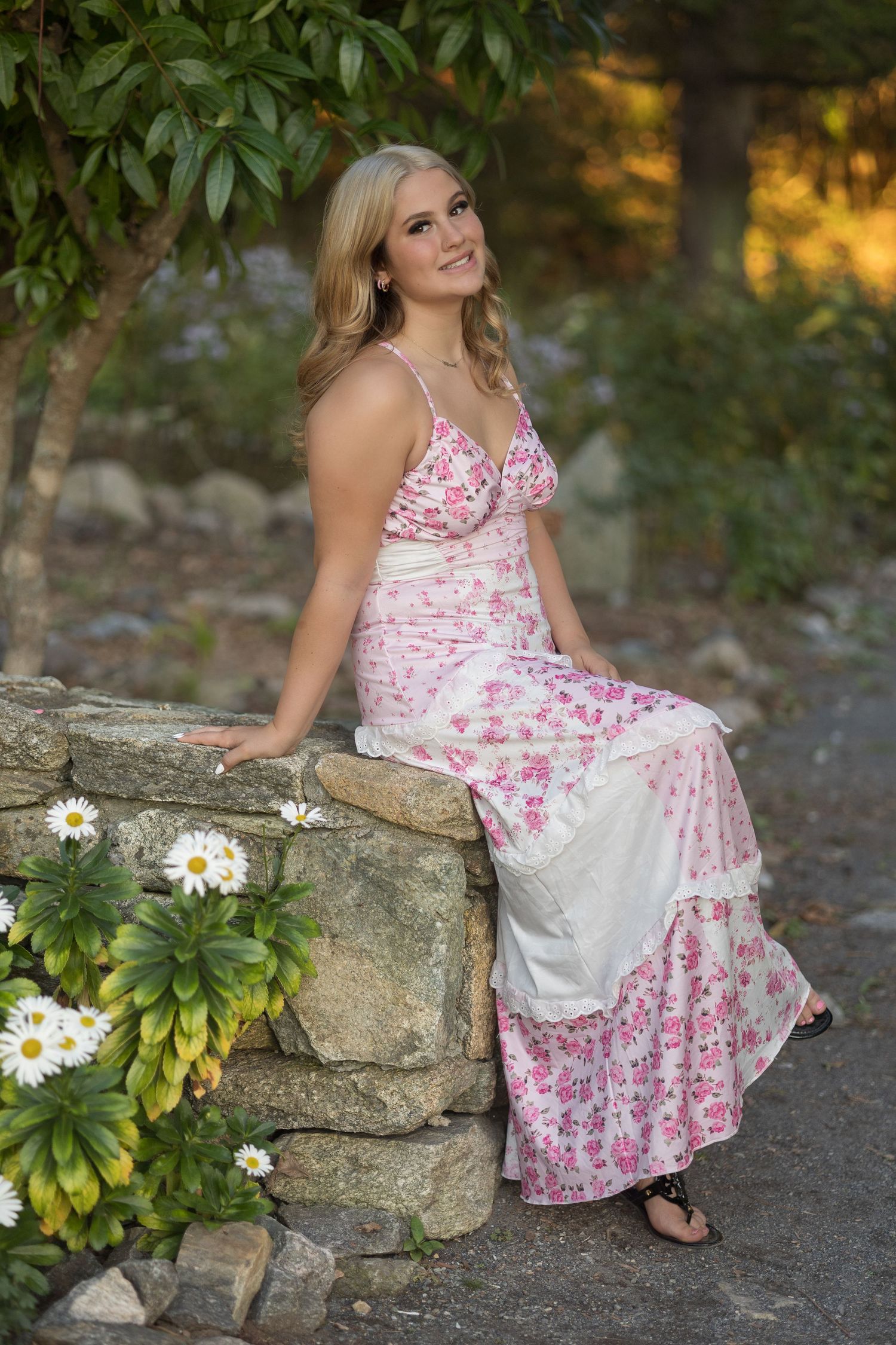 A floral pink and white summer dress is showcased against a rustic stone wall with daisies in the garden setting.