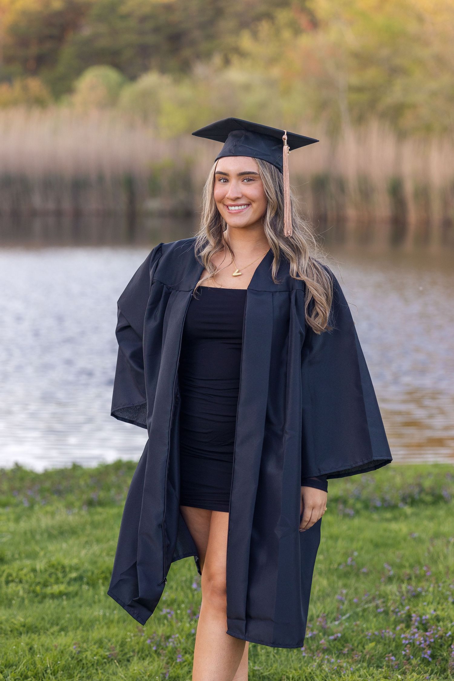 U Mass Dartmouth Graduate wearing black cap and gown poses outdoors near a lake with grassy shoreline during graduation photoshoot.