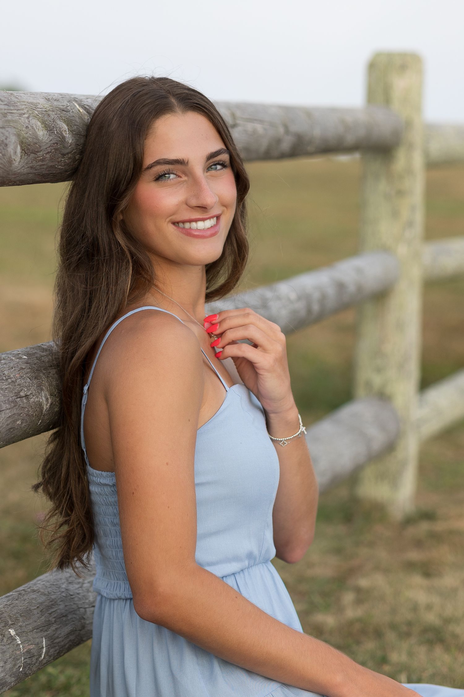A person in a light blue dress poses against a rustic wooden fence in a pastoral outdoor setting.