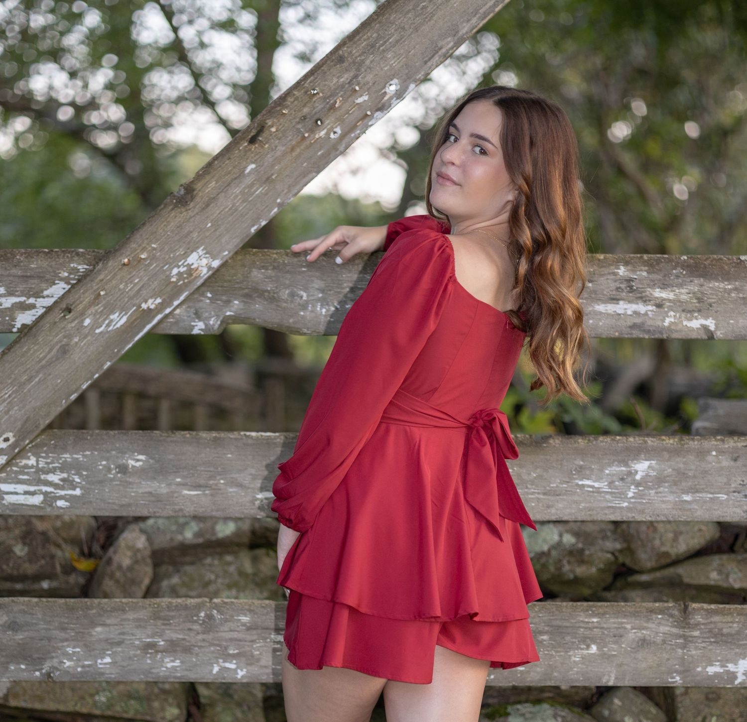 Someone in a flowing red dress poses against a rustic wooden bridge railing in an outdoor setting.
