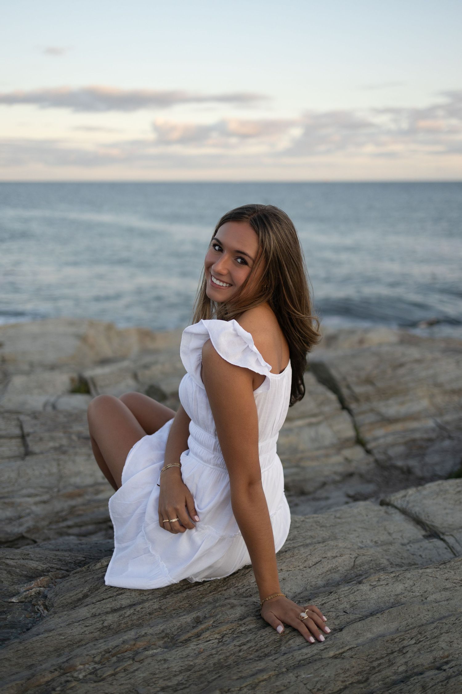 A coastal photoshoot features a white ruffle dress against rocky shoreline at sunset.
