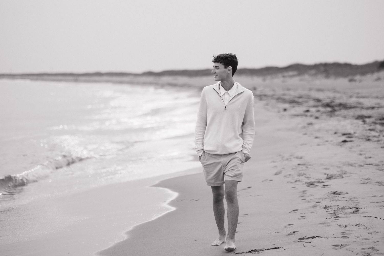 Black and white photo of someone walking alone on a beach wearing a light sweater and shorts.