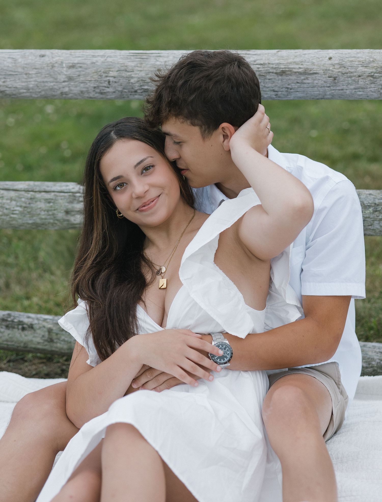 Romantic couple in white outfits sharing intimate moments on a wooden bench in a park setting.