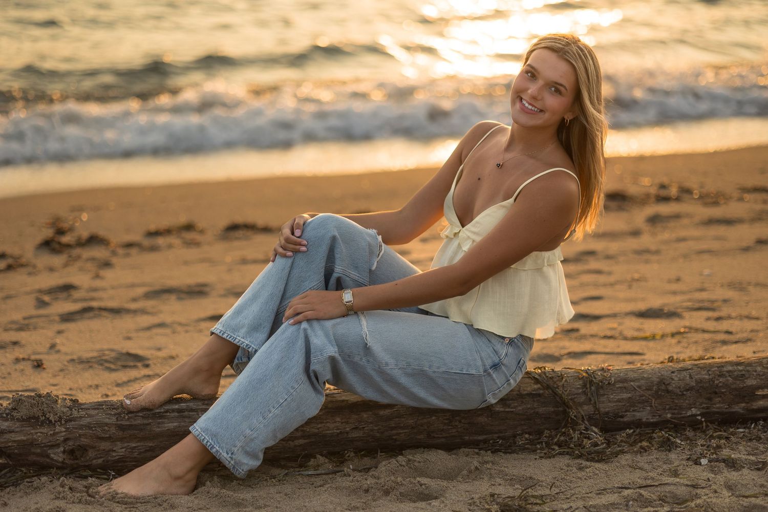 Person in light-colored top and jeans relaxes on driftwood at sunset on a sandy beach.