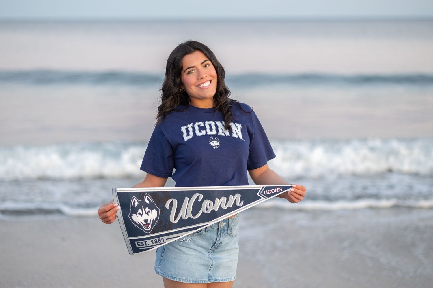Young woman smiling on beach wearing UConn commitment t-shirt and holding UConn pennant flag.