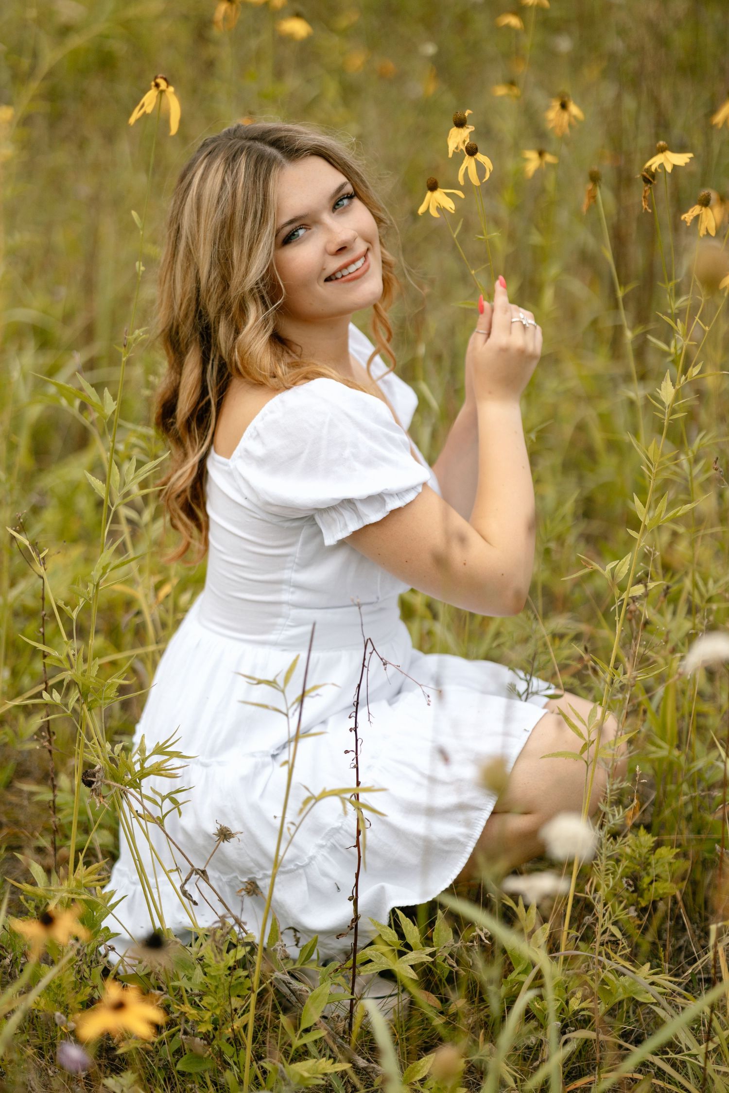 A woman in a white dress poses gracefully among yellow wildflowers in a natural outdoor setting.