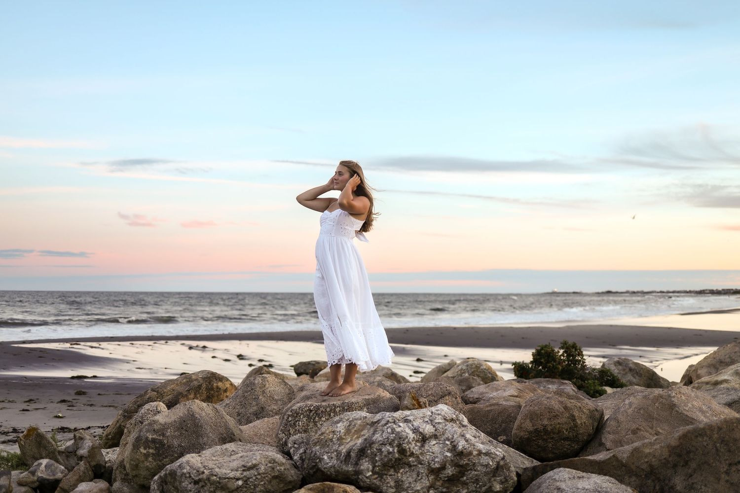 A person in a flowing white dress poses gracefully on coastal rocks at sunset by the ocean.