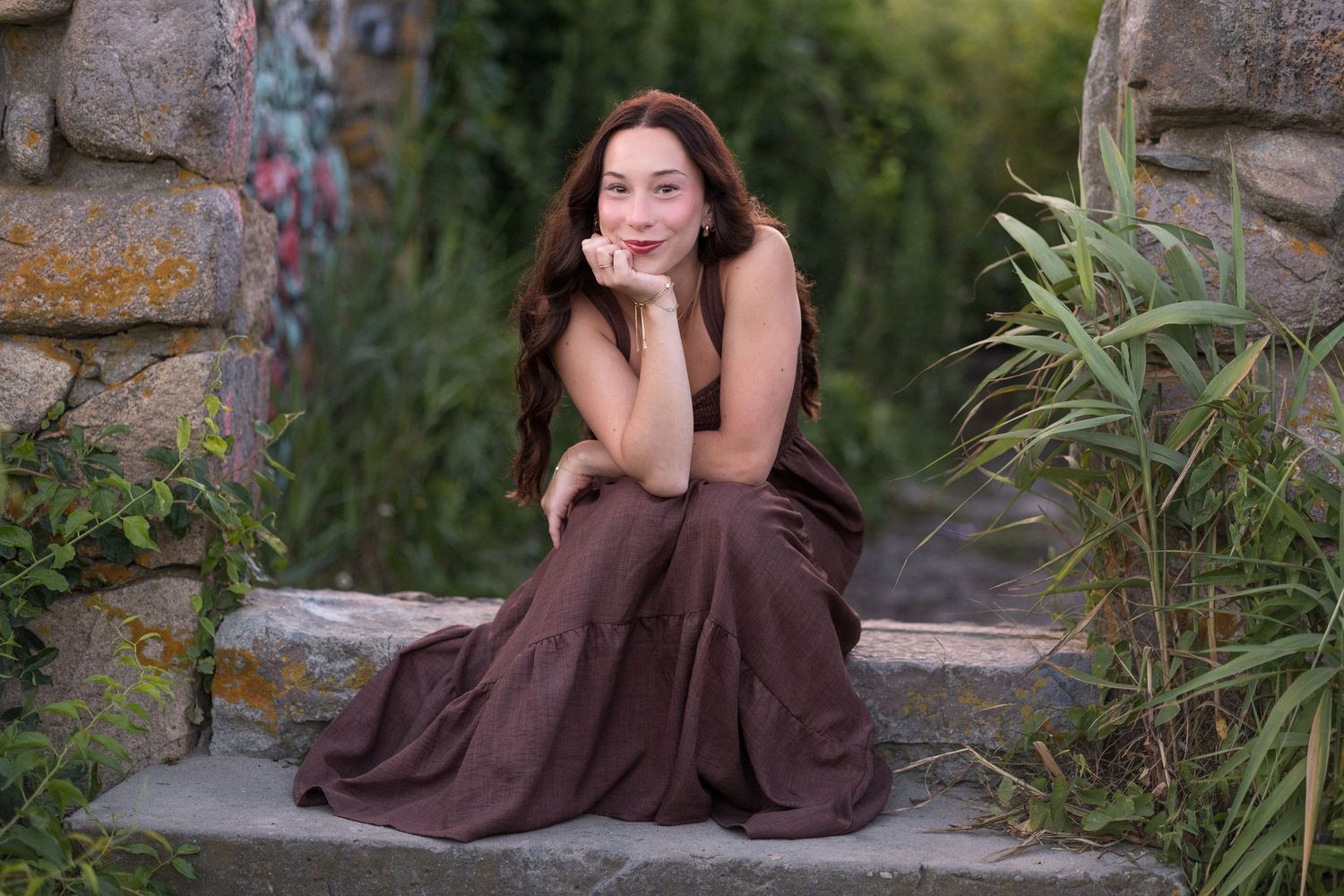 A person in a brown dress poses gracefully on stone steps surrounded by greenery and rustic architecture.