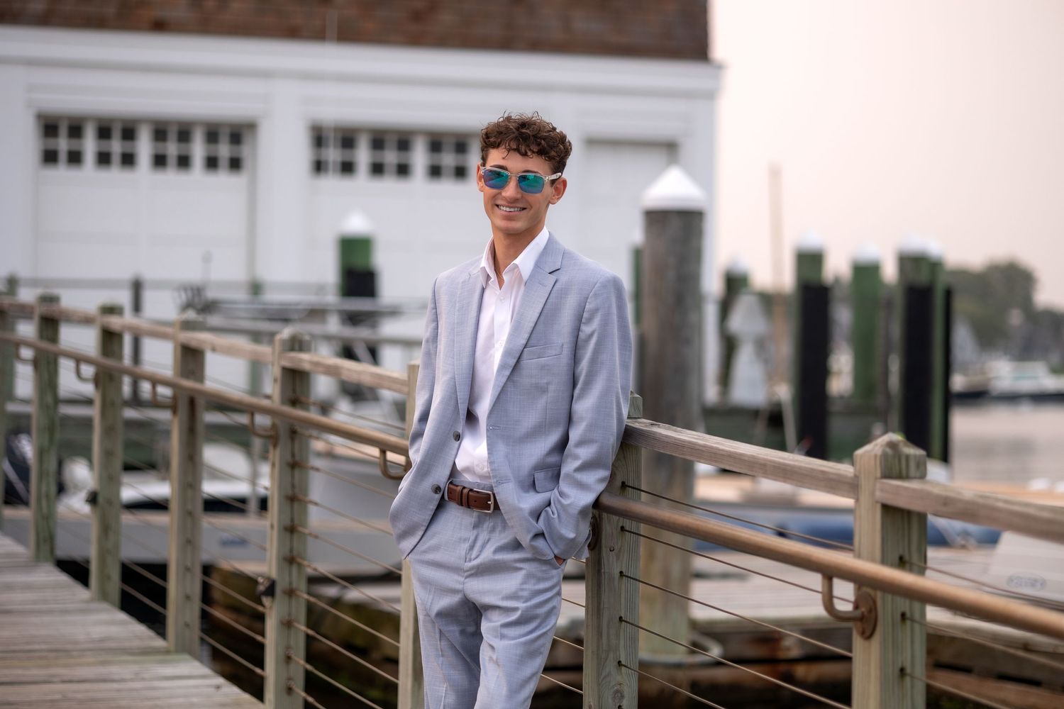 Person in light gray suit stands on coastal boardwalk with boats and water in background.