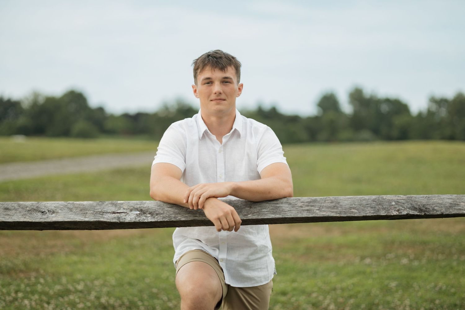 Someone in casual summer attire leans on a wooden fence in an open field.