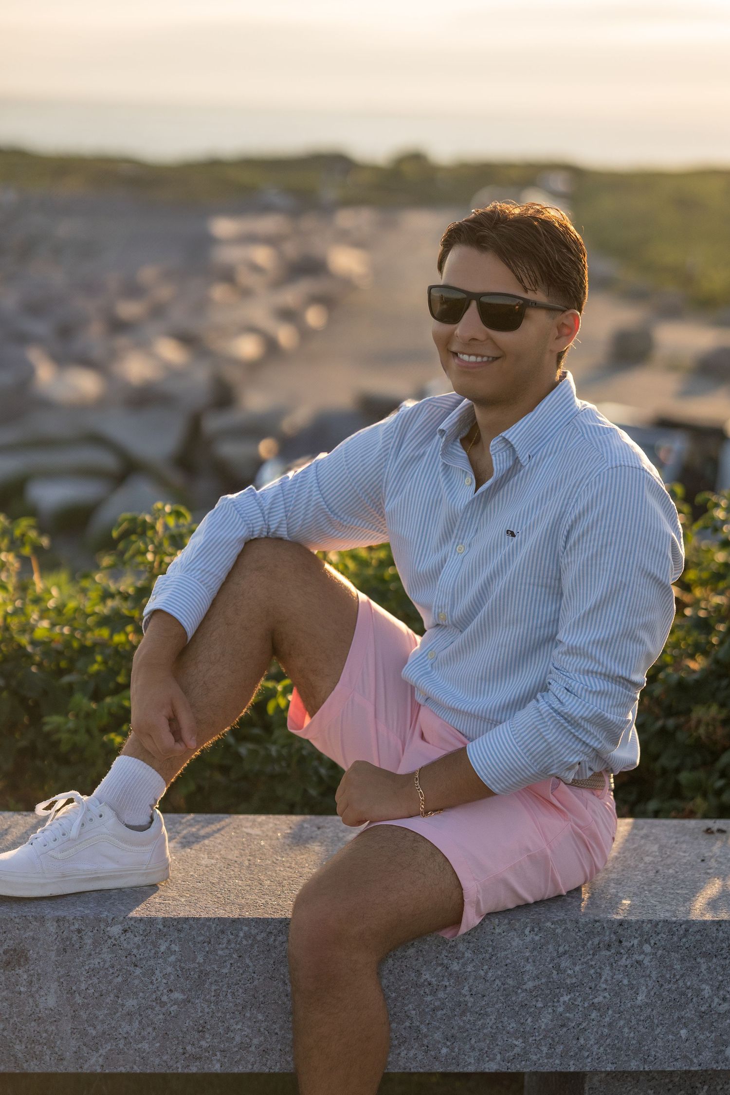 A person in summer attire relaxes on a stone wall at sunset overlooking a coastal parking area.