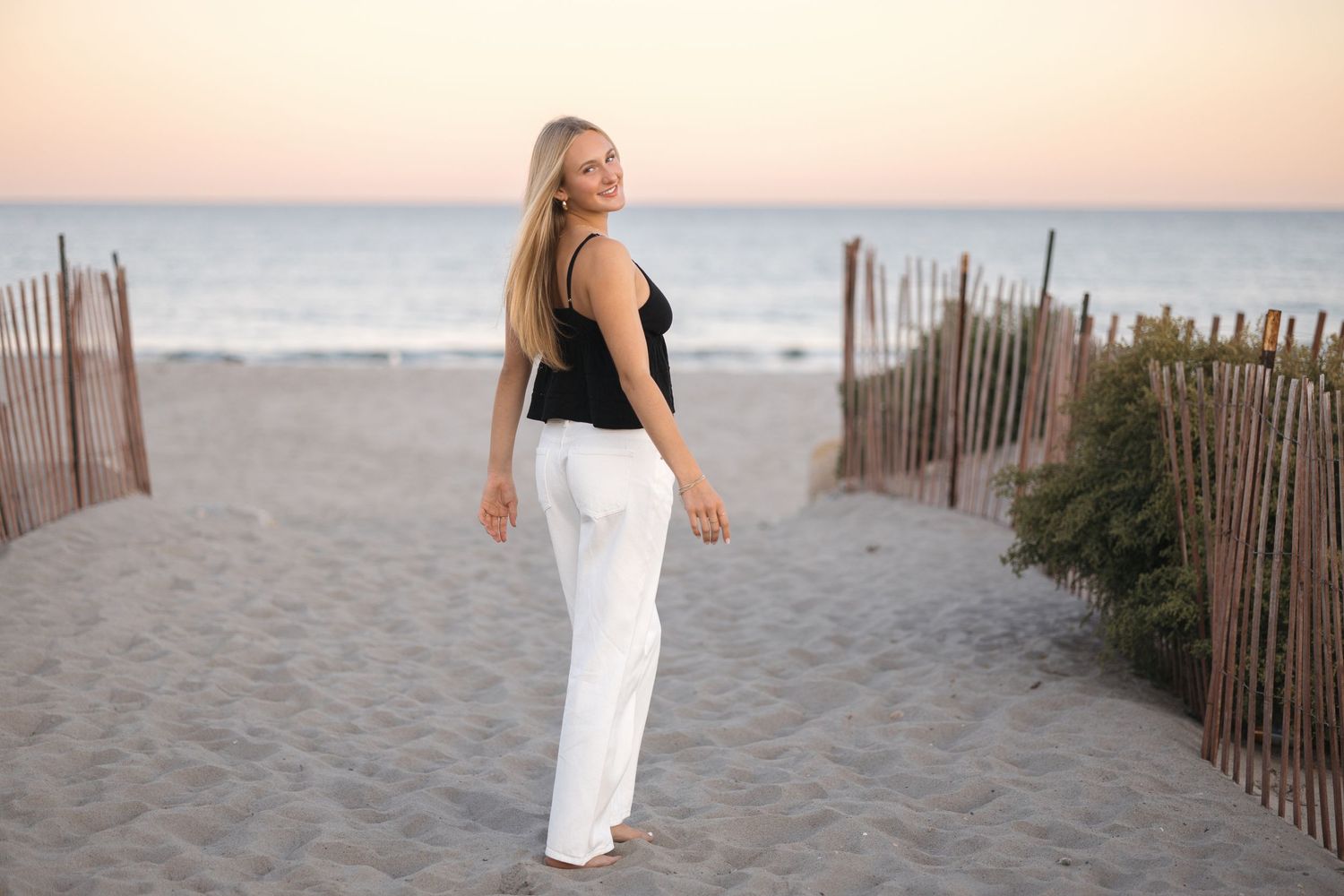 A beachside scene with someone in white pants and black top walking along sandy paths between wooden fences.