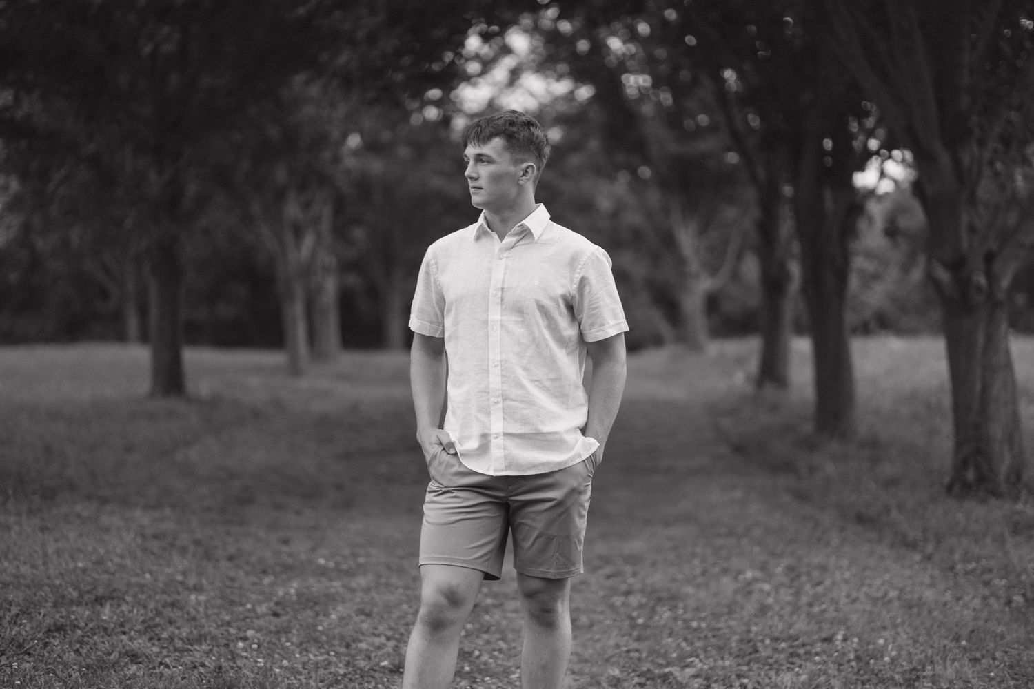 A black and white portrait of someone standing in a park wearing shorts and a short-sleeve shirt.