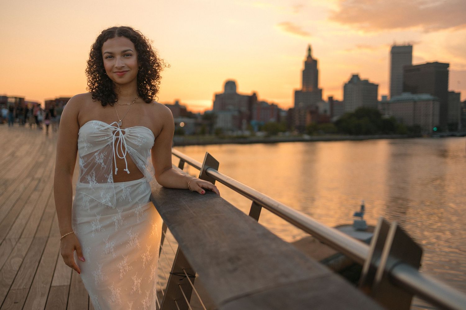A white dress silhouetted against a sunset city skyline along a waterfront deck.