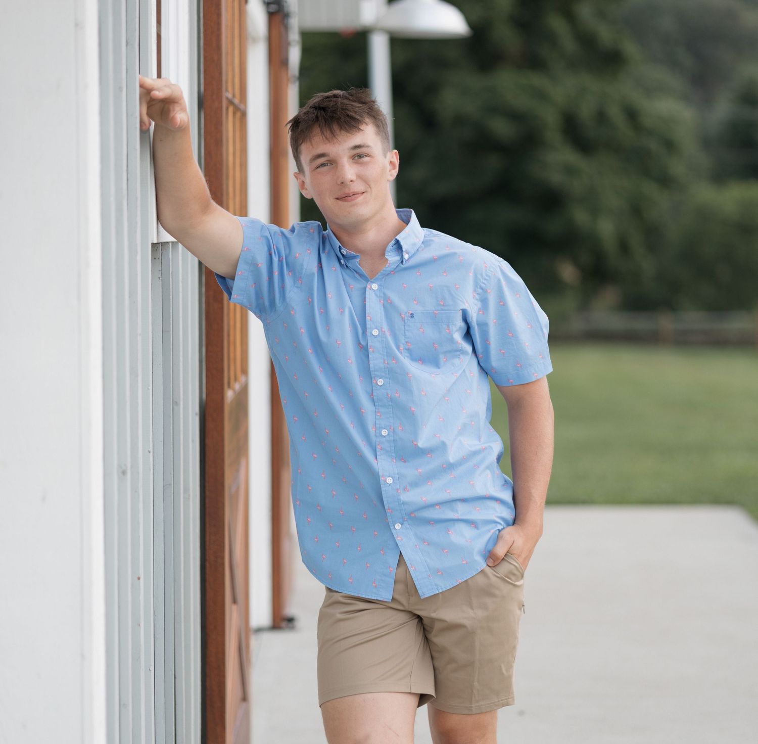 A casual portrait of someone in a light blue shirt and khaki shorts leaning against a white wall.