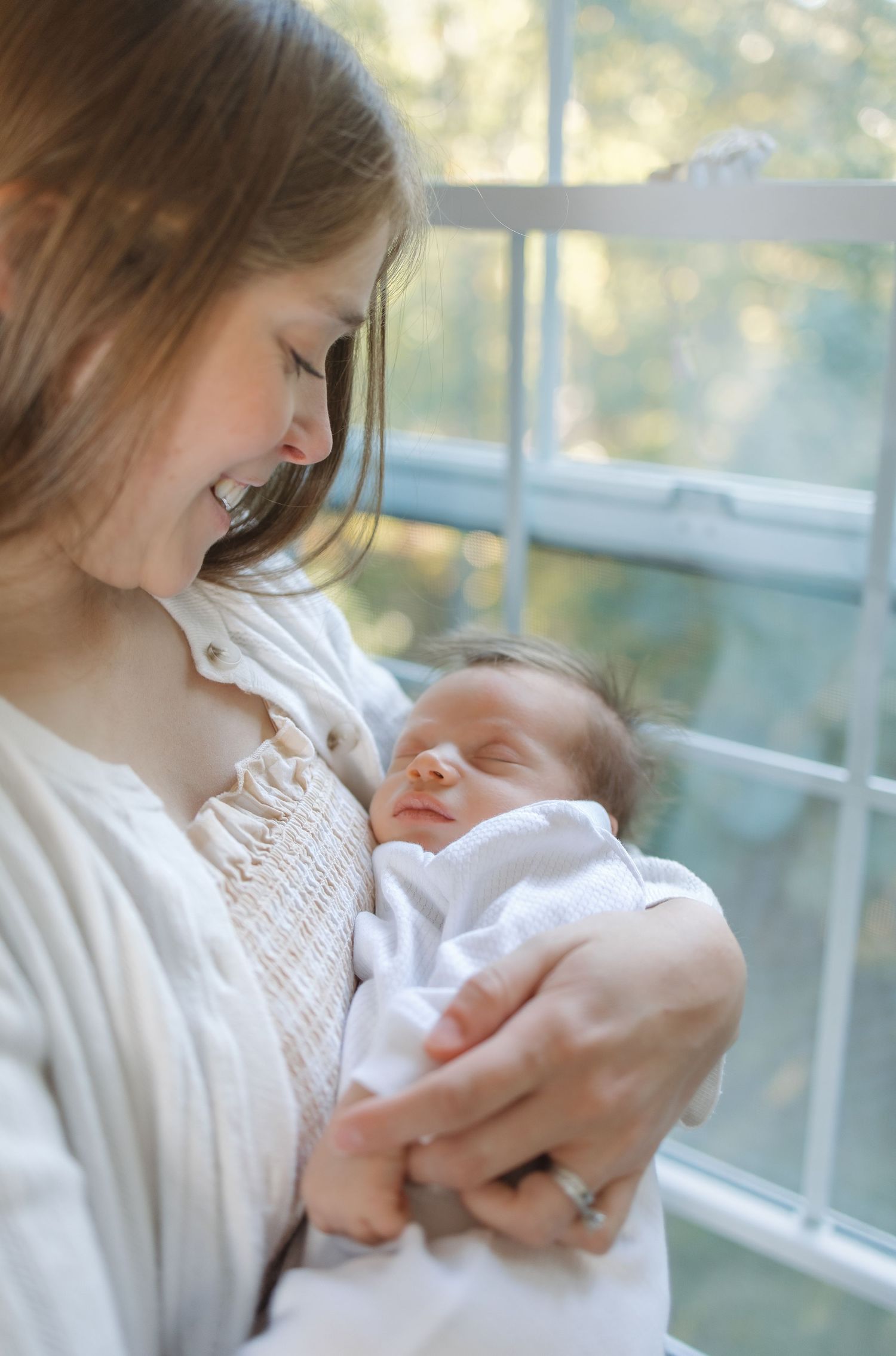 A tender moment between parent and newborn baby swaddled in white, captured in soft natural window light.