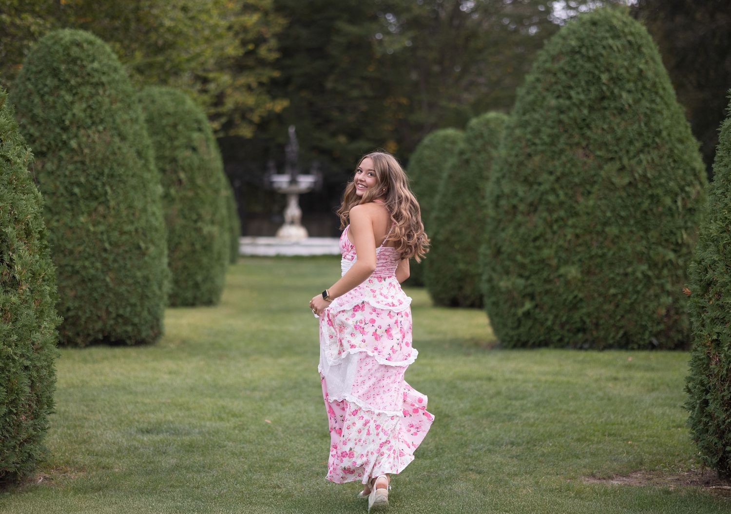 A pink floral dress flows gracefully as someone walks through a formal garden with manicured hedges and fountain.