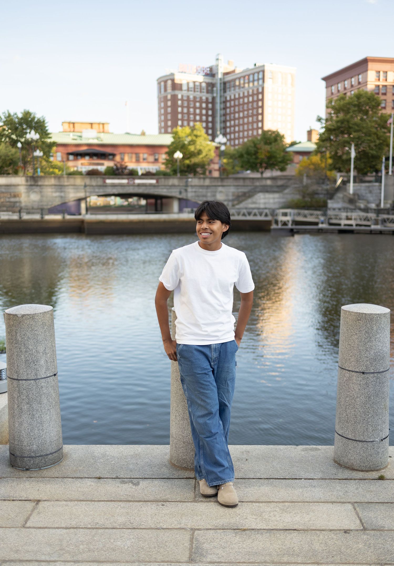 Person poses between concrete pillars along urban waterfront with city buildings in background.