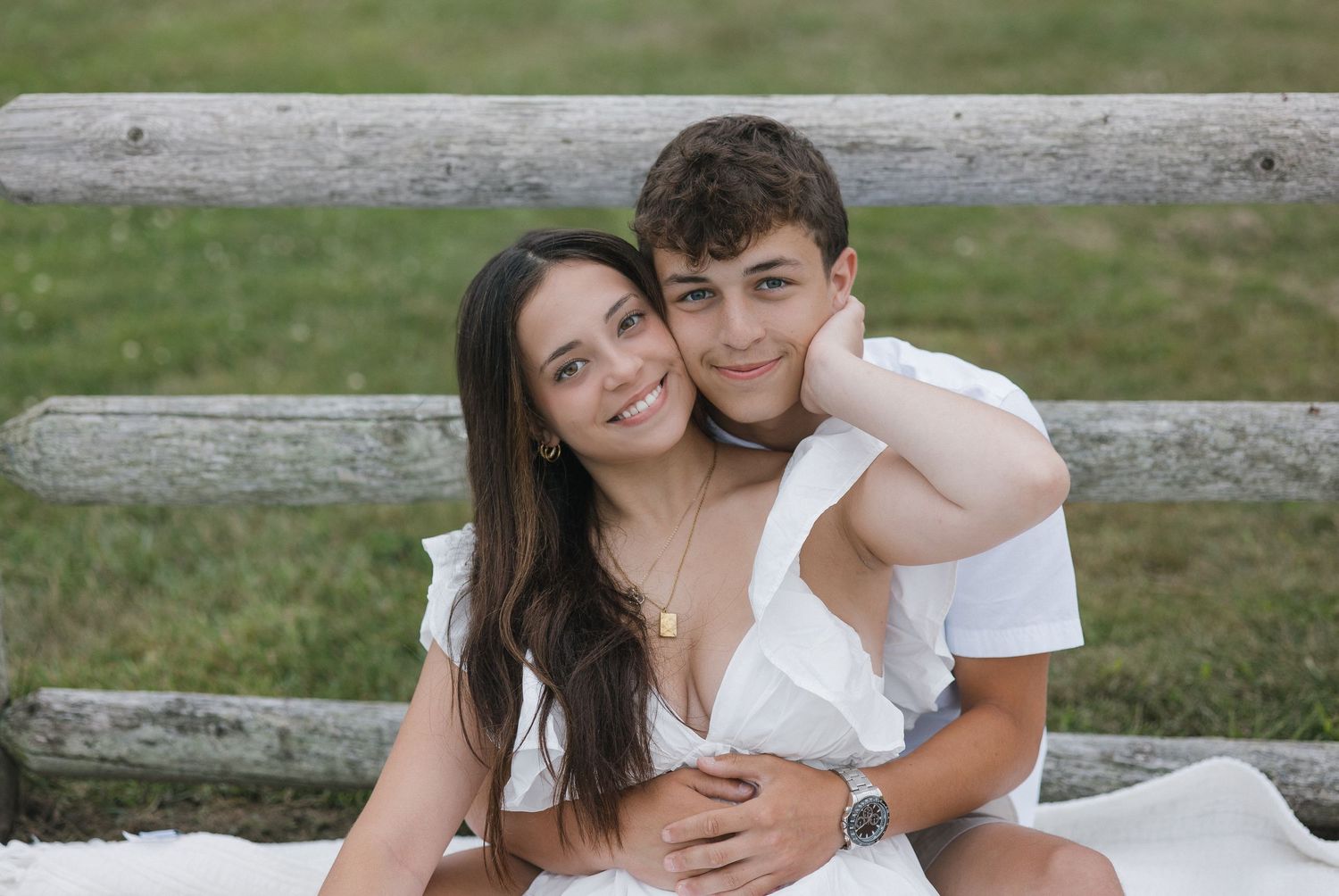 A couple in white outfits embraces on a rustic wooden fence in a pastoral outdoor setting.