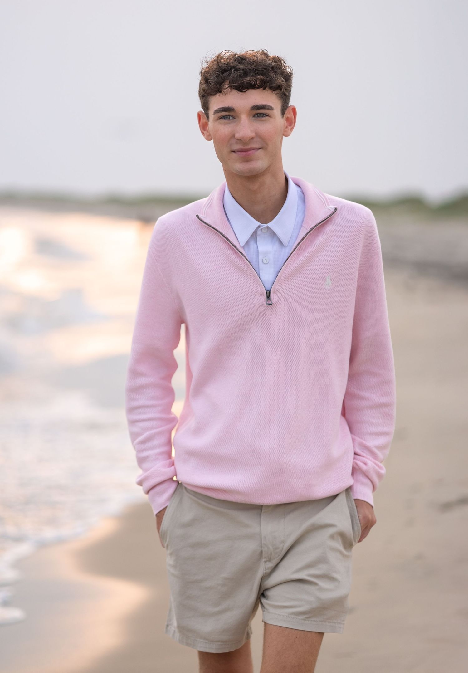 Young person wearing a light pink sweater and khaki shorts poses on a beach at sunset.