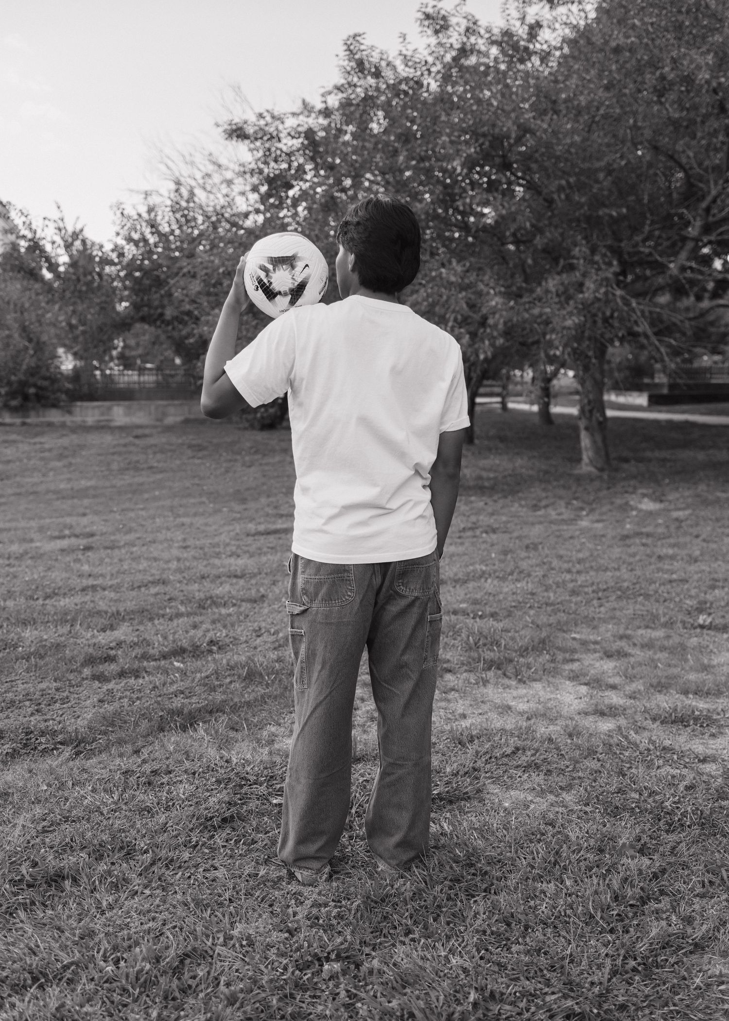 Back view of person in white t-shirt standing in park holding cap.