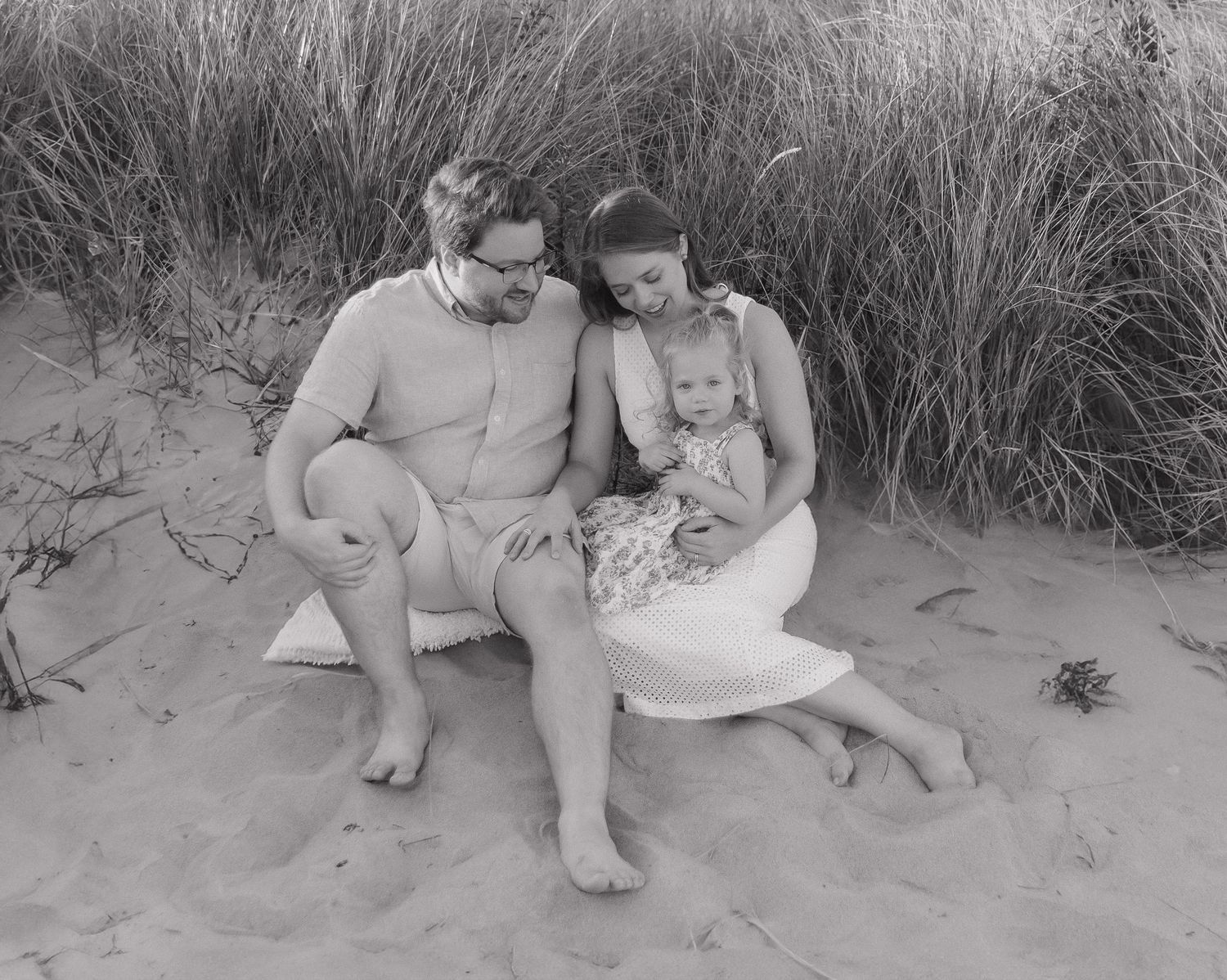 A family sits together in black and white on a sandy beach with tall grass behind them.