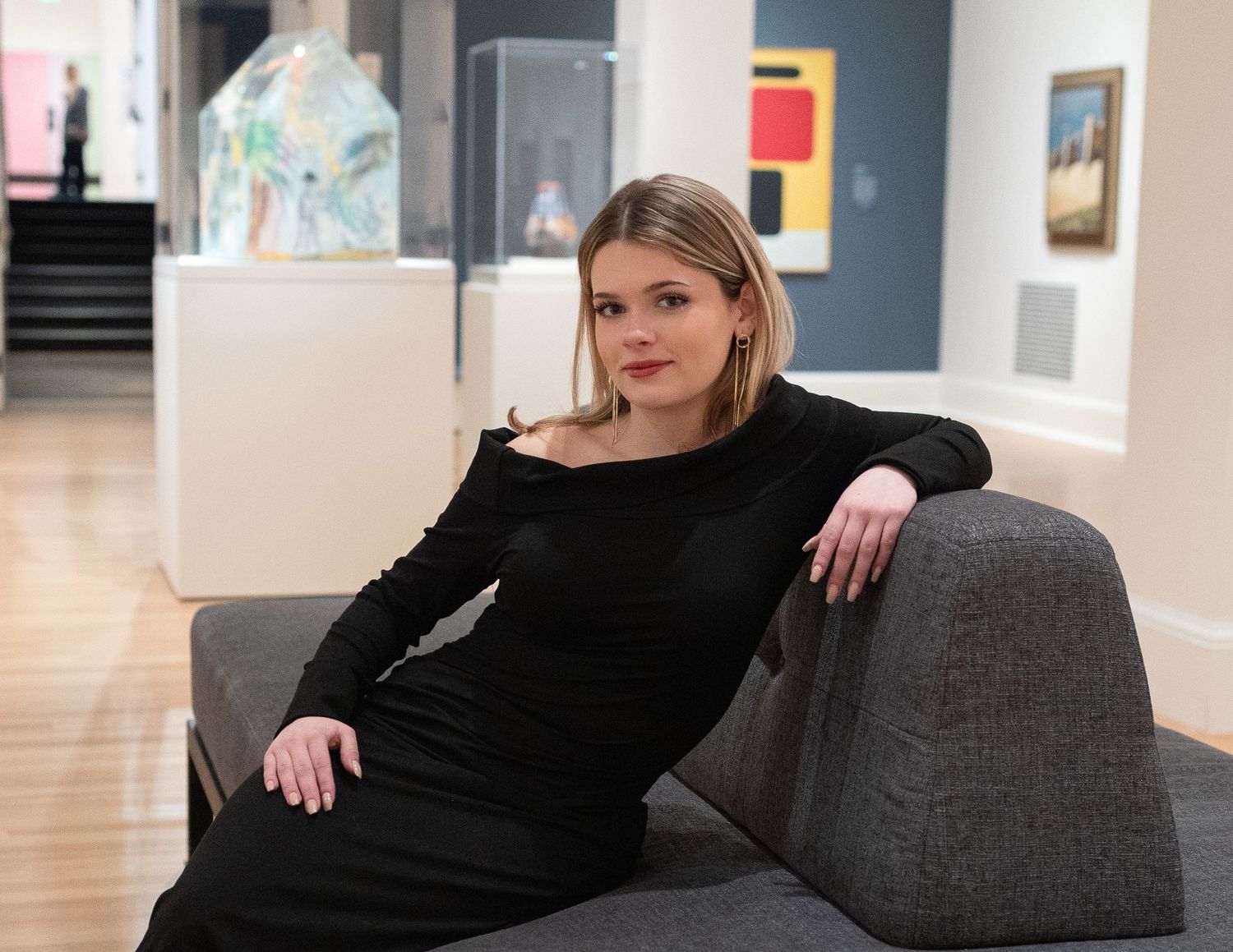 Woman in black dress poses elegantly on grey sofa in bright gallery setting.