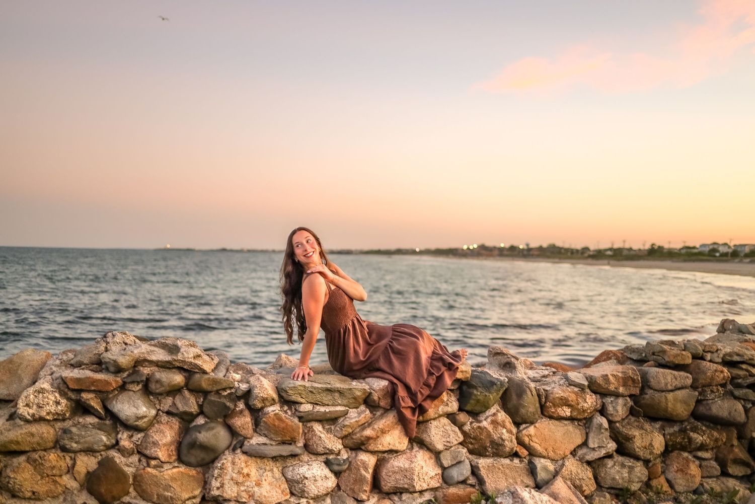 A woman in a flowing dress poses on a stone wall at sunset by the ocean.