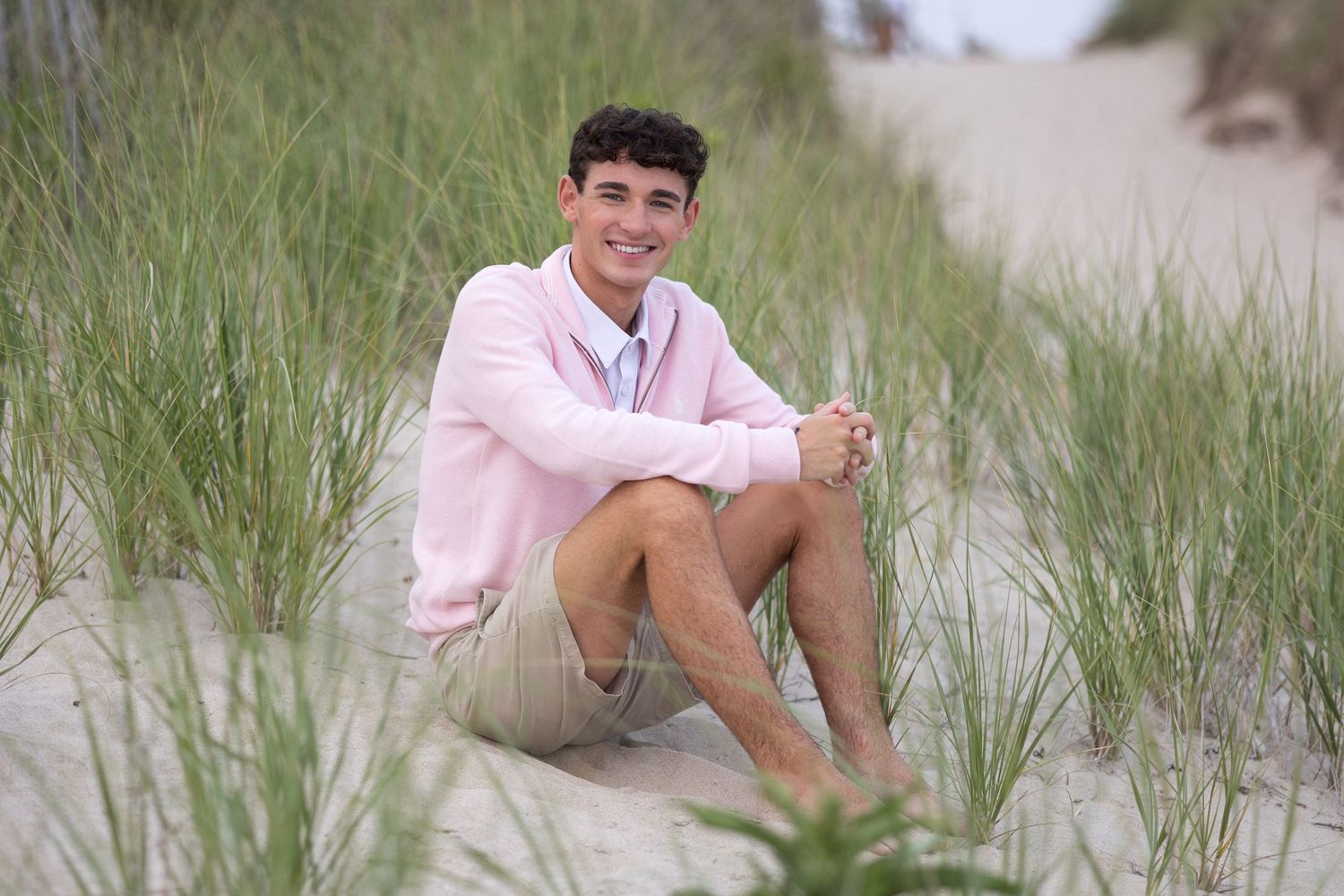 A person in a pink shirt sits casually on sandy dunes surrounded by tall beach grass on a cloudy day.