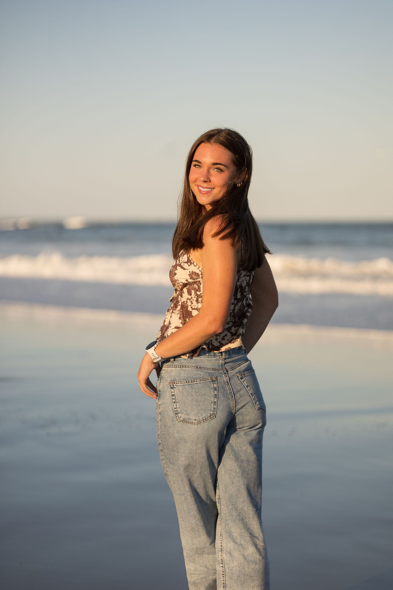 A beachside photoshoot featuring casual denim and floral top against the ocean backdrop at sunset.