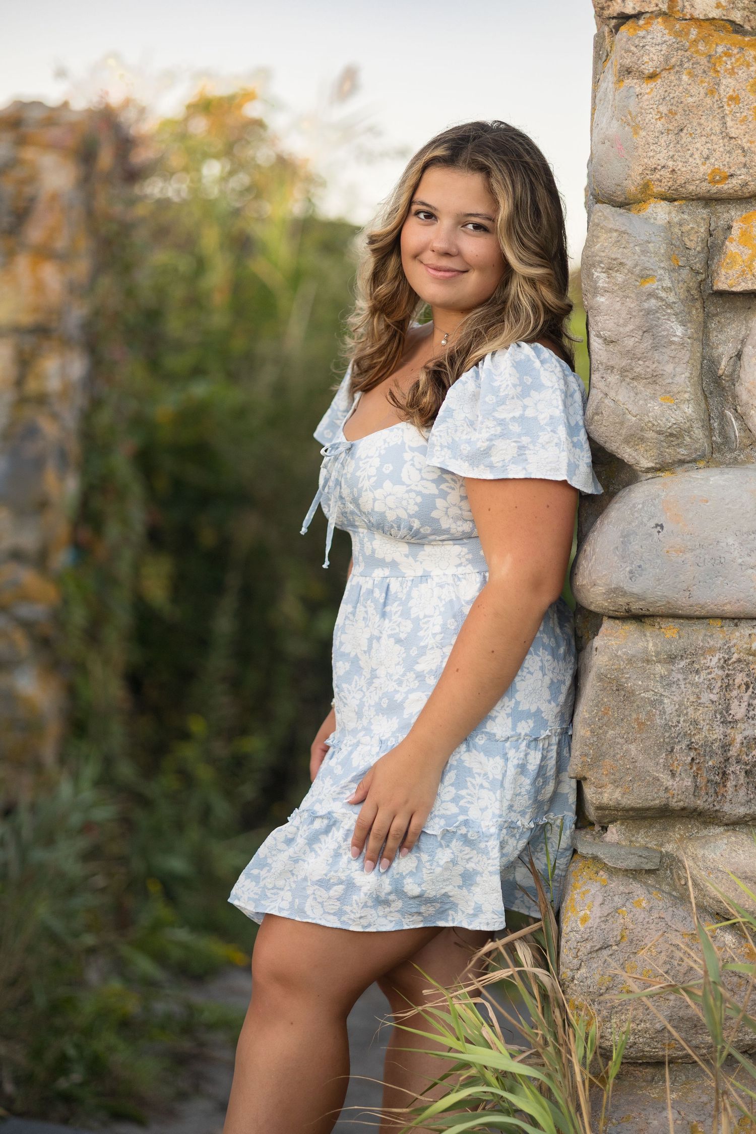 A young girl in a blue and white floral dress poses outdoors near a stone wall during a summer photo session.