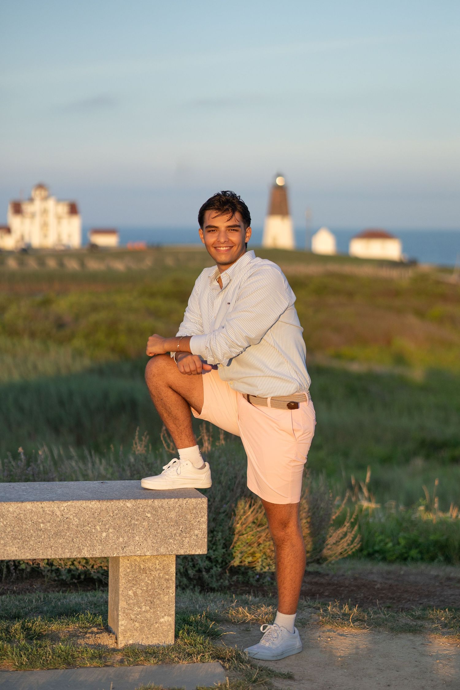 A person in casual attire sits on a concrete block at sunset with coastal scenery in the background.