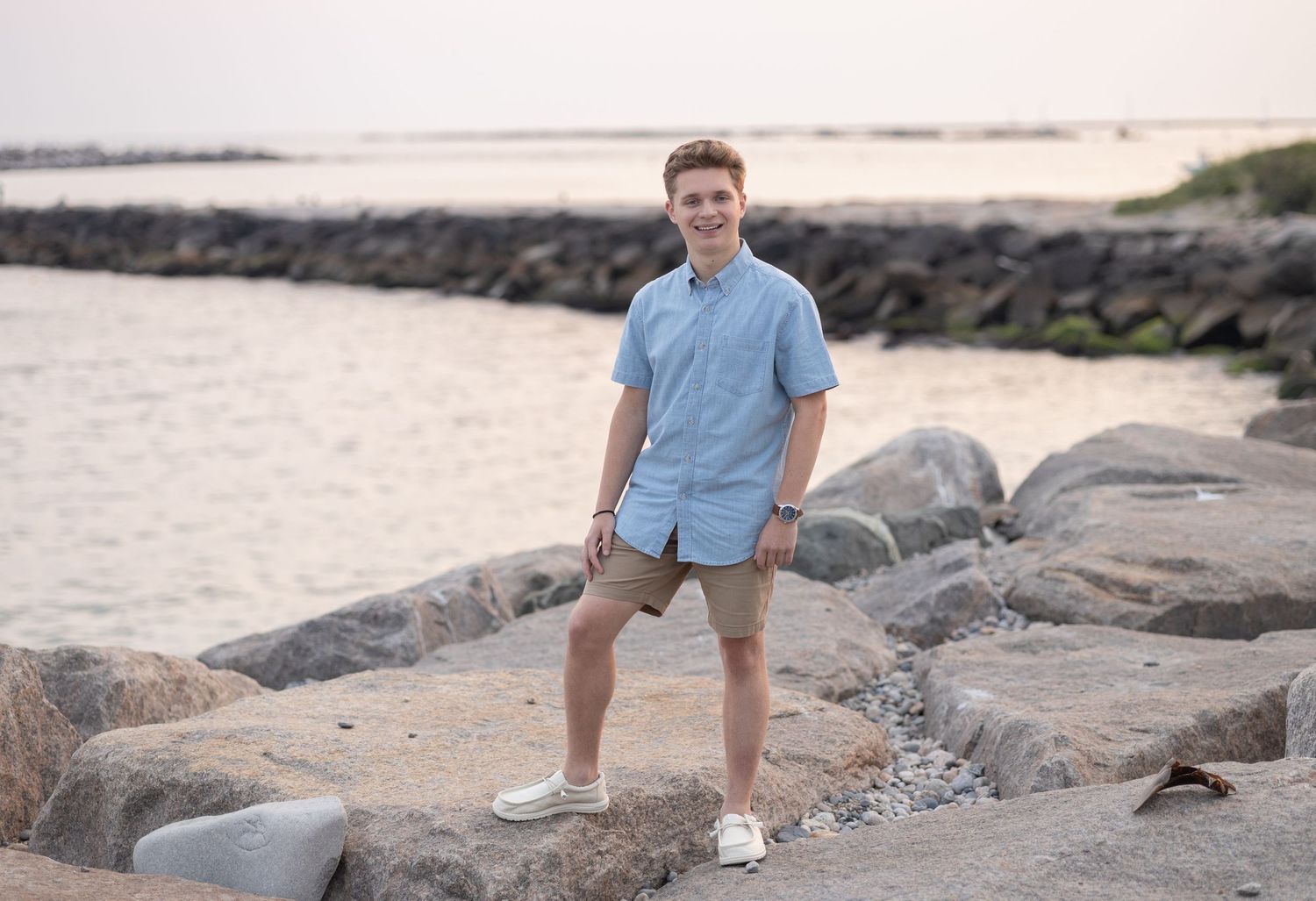 A casual pose on coastal rocks with ocean and jetty in the background.
