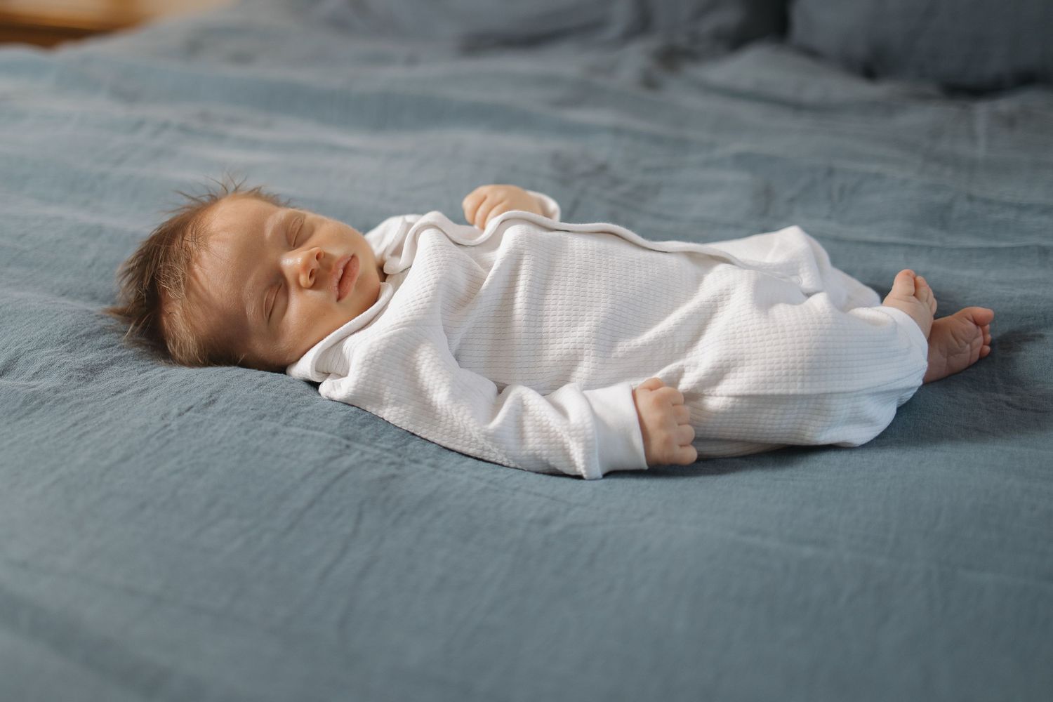 A newborn baby wearing a white onesie sleeps peacefully on soft grey bedding in a side-lying position.