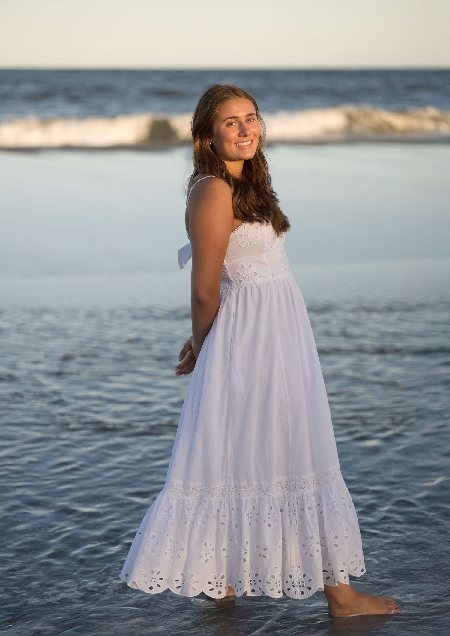 A person in a white eyelet lace maxi dress poses at sunset on the beach with ocean waves in the background.