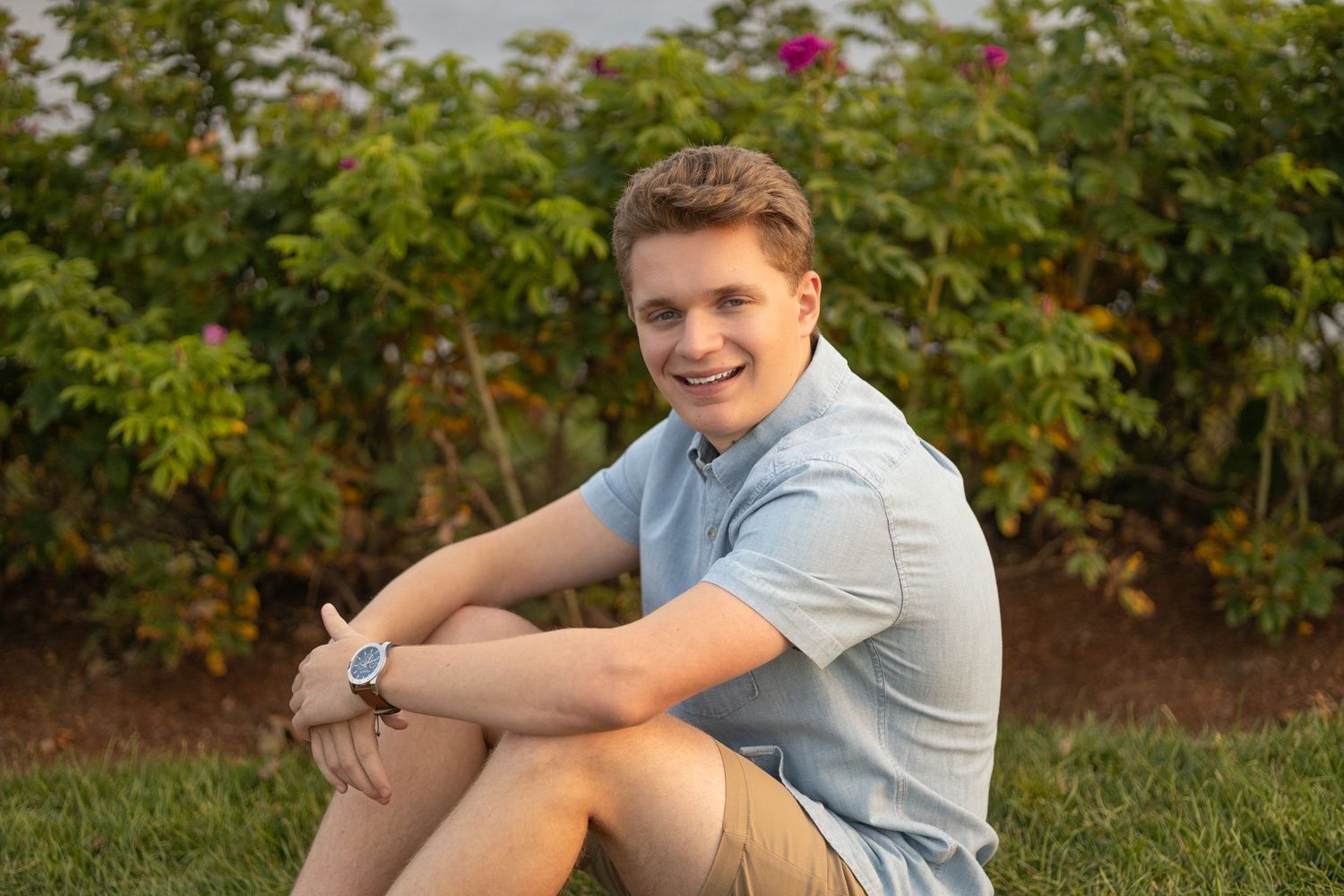 Someone sits casually on grass with blooming rose bushes in the background.