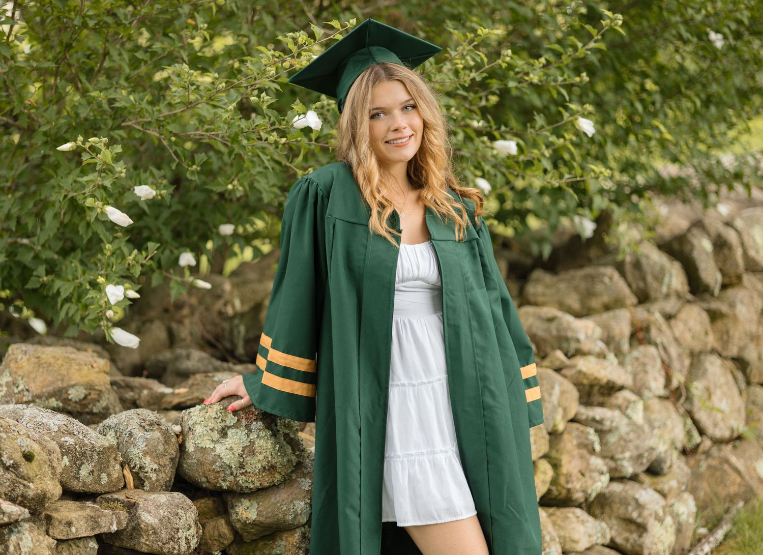 A graduate stands proudly in green commencement robes against a rustic stone wall background.