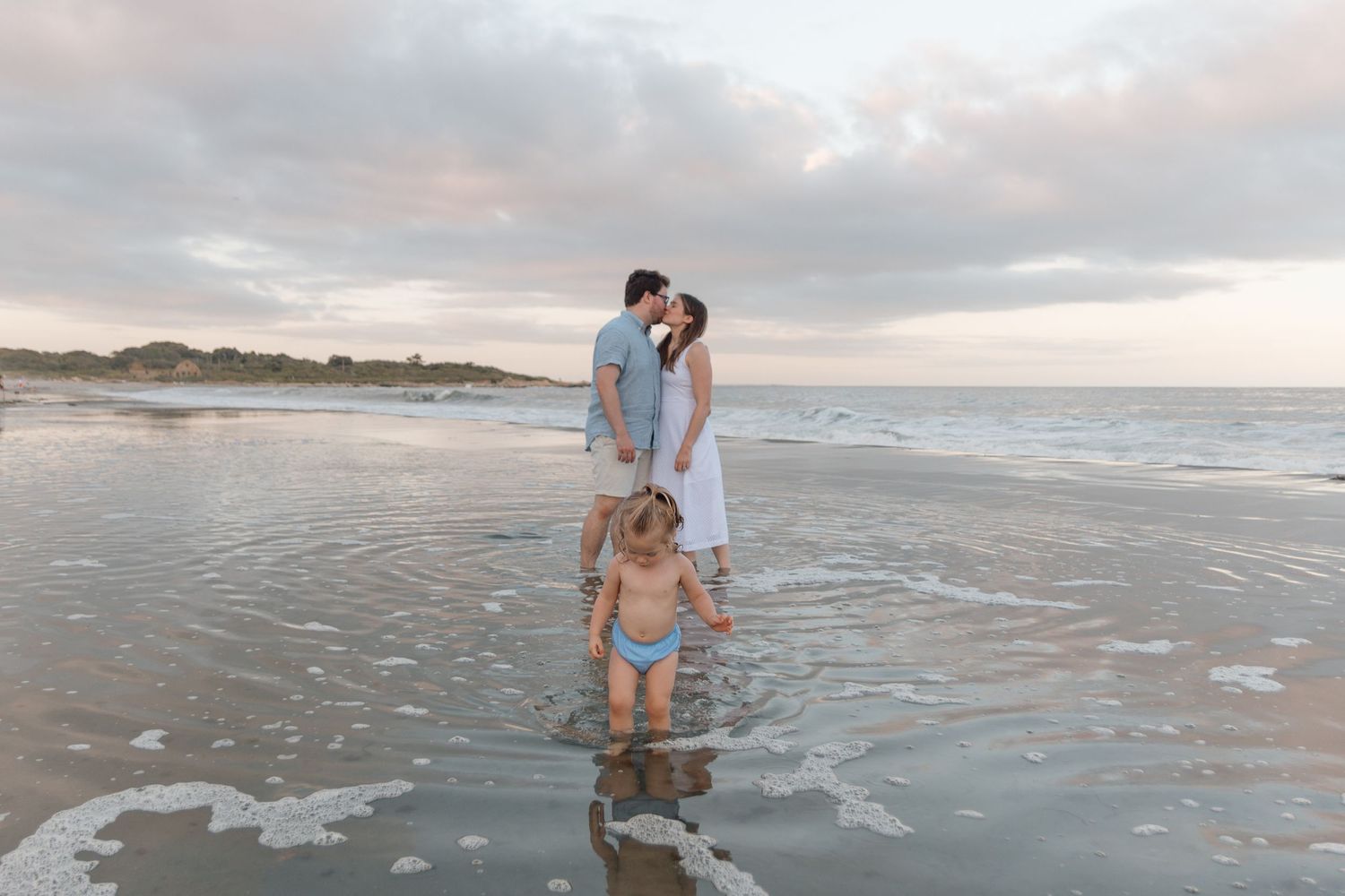Family enjoying a serene sunset moment together on a beach with wet sand and cloudy skies.