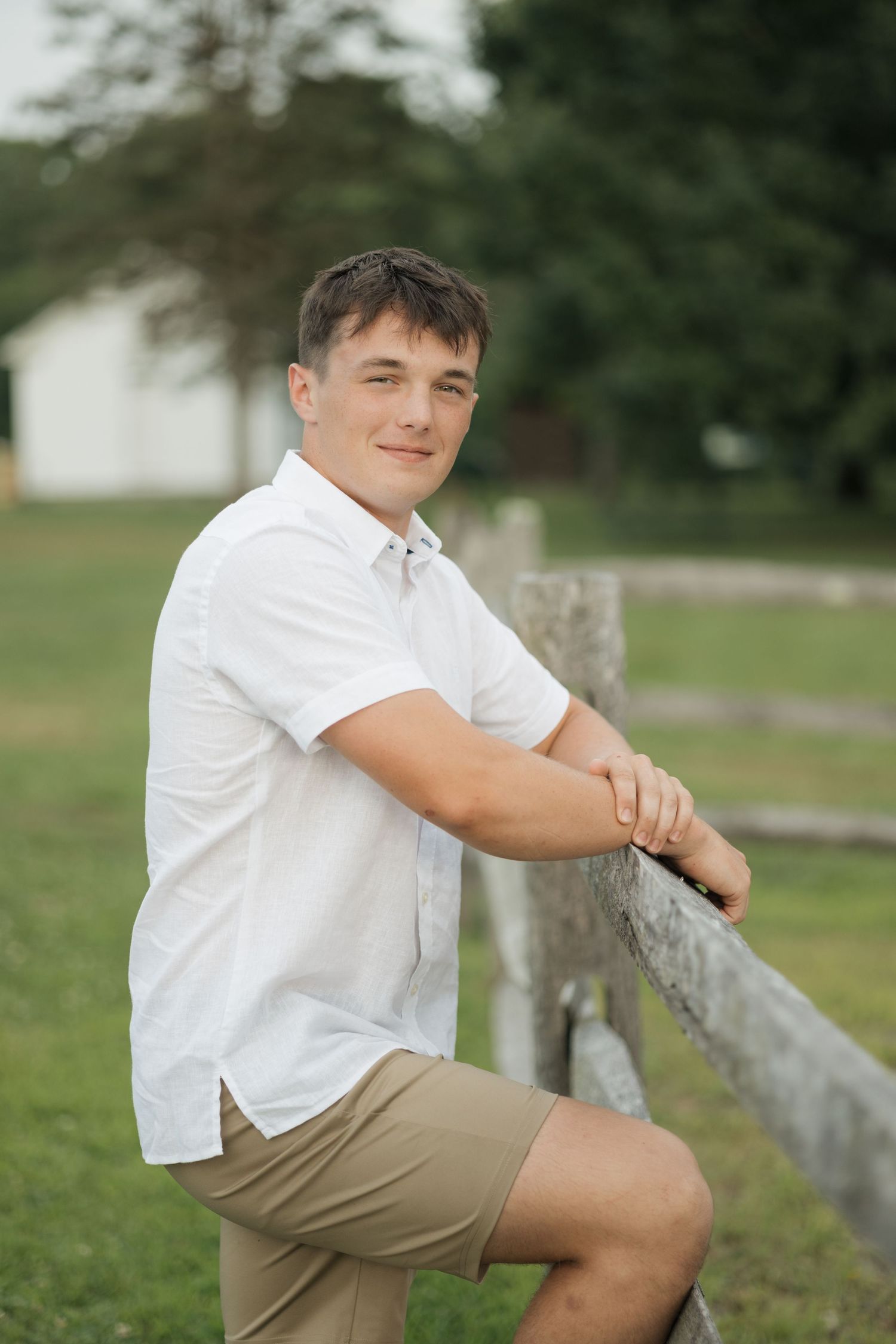 Person in white polo shirt leaning on wooden fence in countryside setting.