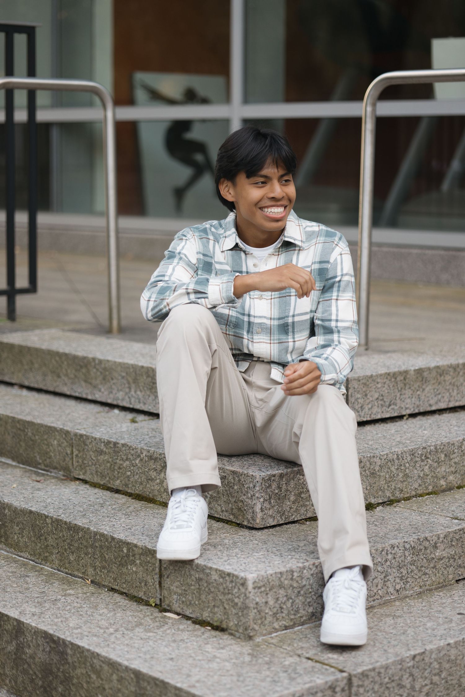 Young person in plaid shirt and khaki pants sits casually on concrete steps wearing white sneakers.